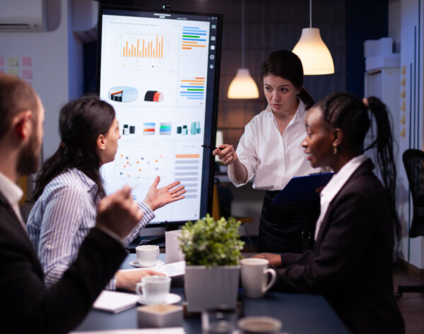 Multi-ethnic businesspeople discussing financial company solution sitting at conference table in meeting room. Diverse teamwork overworking at management strategy using tv late at night.